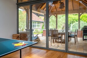 Ein Zimmer mit Tischtennisplatte, einem Essbereich mit Stühlen und Tisch sowie Blick auf einen Garten durch große Glastüren.