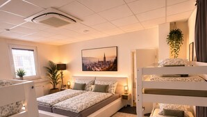 Et hotelværelse med køjesenge, en stor seng, et bybillede som maleri, en potteplante og et vindue med persienner.
