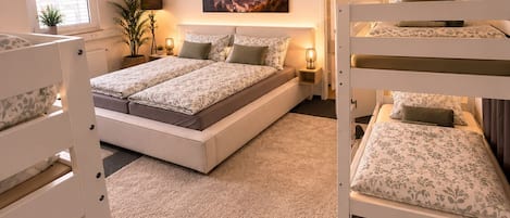Habitación de hotel con literas, una cama grande, un cuadro de un paisaje urbano, una planta en maceta y una ventana con persianas.