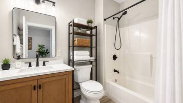 A bathroom with a white vanity, a toilet, a shower with a black hose, and a shelf with towels and a plant.