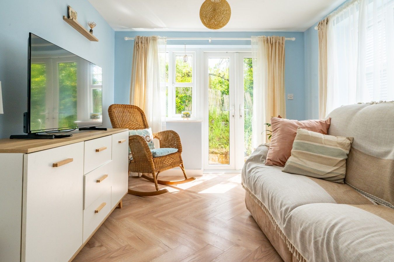 Ein Zimmer mit Fernseher auf einem Holzschrank, einem Korbstuhl, einer Couch mit Kissen und Blick ins Grüne durch eine Glastür.