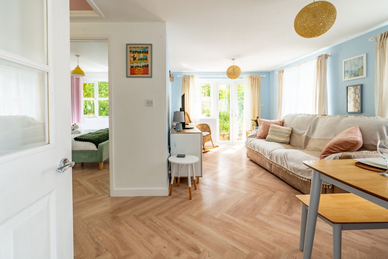 A well-lit room with a wooden floor, a sofa, a dining table, and a chair. There is a door leading to another room.