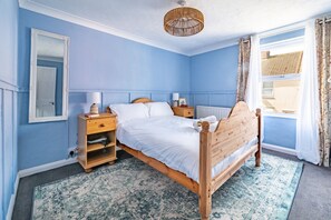 Ein Schlafzimmer mit einem hölzernen Bett, einem Nachttisch, einem Spiegel, einem Fenster mit Vorhängen und einer Deckenleuchte.