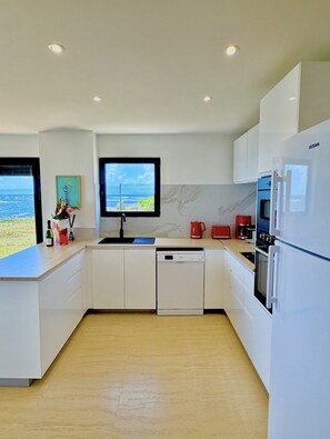 A modern kitchen with white cabinetry, a built-in dishwasher, a large refrigerator, and a flat-screen TV mounted above the sink. - SEA VIEW TINTAMARRE Villa – Brand-new waterfront villa in Oyster Pond (Oyster Pond)