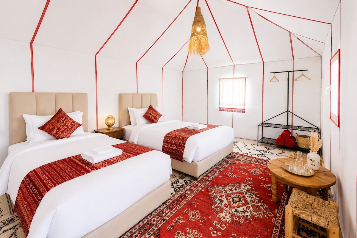 A hotel room with two beds, a red patterned rug, a wooden side table, and a hanging light fixture.