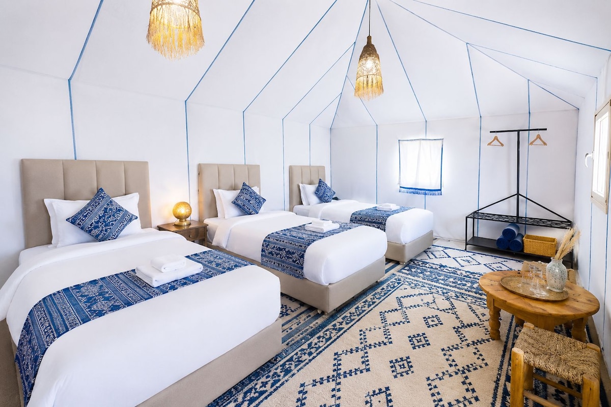 A hotel room with three beds, a small table, a hanging light fixture, and a patterned rug.