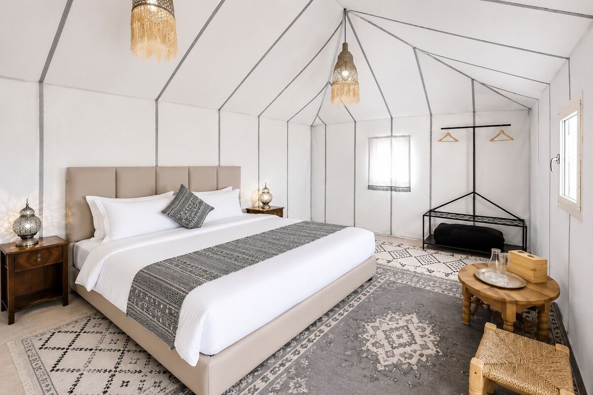 A modern bedroom with a large bed, a nightstand, a bedside table, a hanging light fixture, a window, and a wooden bench.