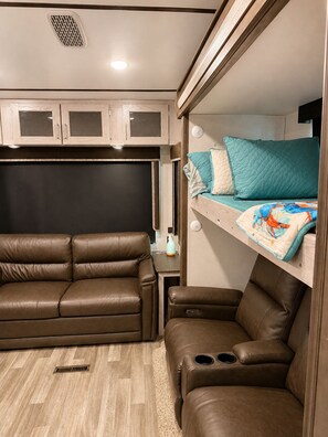Een compact RV-interieur met een bank, een zitgedeelte beneden en een stapelbed met kussens.