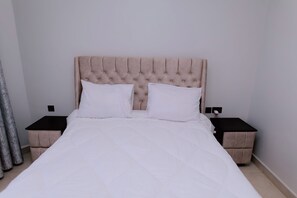A bed with a tufted headboard, two white pillows, and a white duvet. There are two bedside tables with black tops and beige sides. - Luxury 3-Bedroom Suite · Gishushu, Kigali (Kigali)