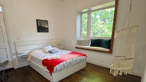 Ein Schlafzimmer mit Bett, Fenster mit Blick, einem Makramee-Stuhl und einem Schreibtisch.