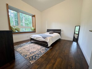 A bedroom with a bed, a window with a view of trees, a dark wooden floor, and a large mirror. - Branch & Balsam House
Modern Forest Retreat (Huntsville)