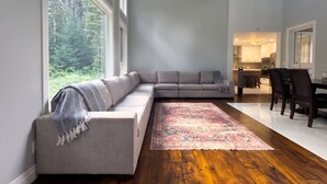 Ein modernes Wohnzimmer mit einem grauen Ecksofa, einem gemusterten Teppich und Blick auf einen Wald durch große Fenster.
