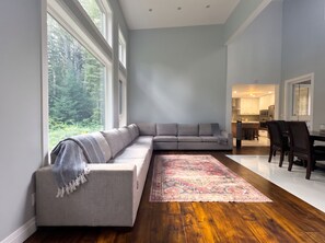 Een moderne woonkamer met een grijze modulaire bank, een getextureerd vloerkleed en uitzicht op een bos door grote ramen.
