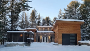 Ein modernes Blockhaus mit einem zentralen Eingang, umgeben von schneebedecktem Boden und Bäumen.