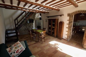 Ein rustikaler Innenraum mit Kamin aus Stein, einer Holztreppe und einem Essbereich mit Tisch und Stühlen.