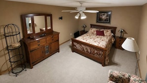 Een slaapkamer met een bed, een dressoir, een spiegel, een plafondventilator, een lamp en een stoel.