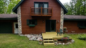Een houten chalet met een balkon, een voordeur en een klein terras met treden naar een tafel en stoelen.