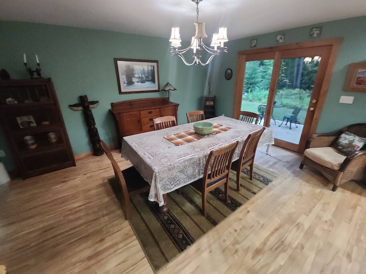 Un coin repas avec une table dressée pour quatre personnes, entourée de chaises en bois, d’un lustre et offrant une vue sur l’extérieur à travers une porte vitrée.