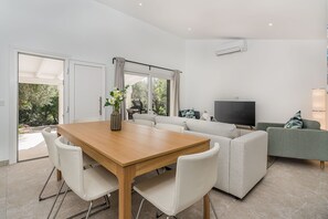 A modern living room with a wooden dining table, white chairs, a grey sofa, and a television. - Premium Pollensa Country Villa for 6 (Pollença)