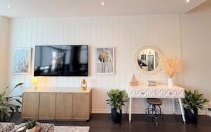 Een moderne woonkamer met een flatscreen televisie, een ronde spiegel, een klein tafeltje met een krukje en decoratieve planten.