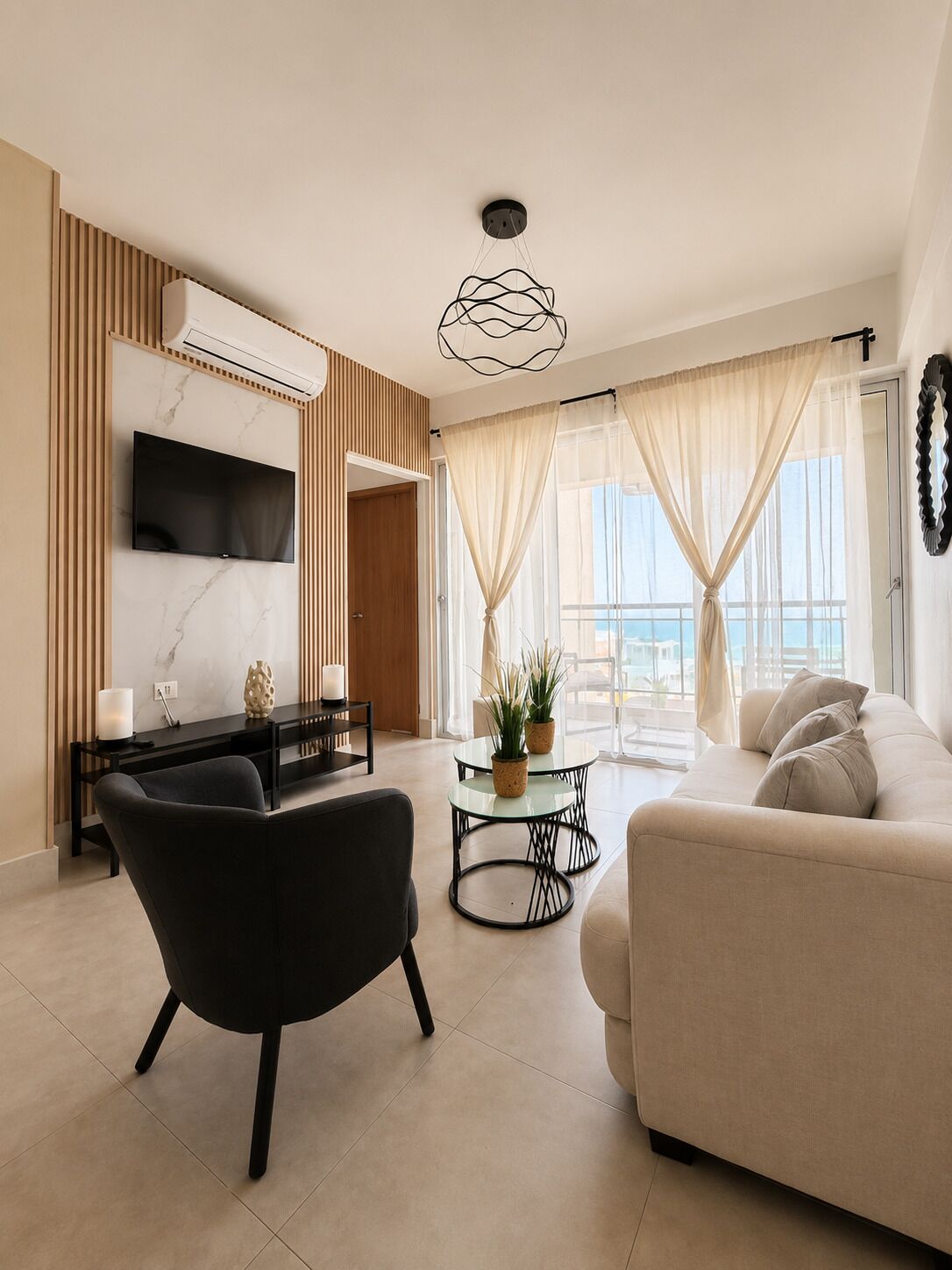 Un moderno salón con televisor de pantalla plana, una silla negra, un sofá beige, una mesa de centro de vidrio con plantas y vistas al mar a través de cortinas traslúcidas.
