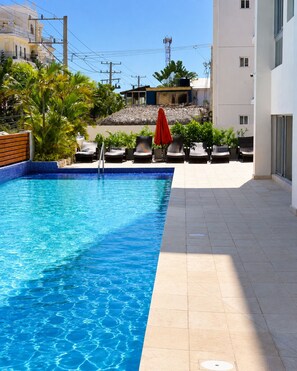 A swimming pool with lounge chairs and umbrellas, surrounded by buildings and vegetation. - Bright 2-bedroom apartment with AC in tranquil Boca Chica (Boca Chica)