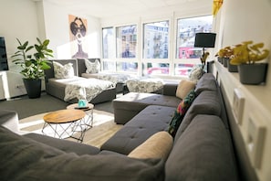 Ein modernes Wohnzimmer mit einem grauen Ecksofa, einem runden Couchtisch und einem großen Fenster mit Blick auf Gebäude.