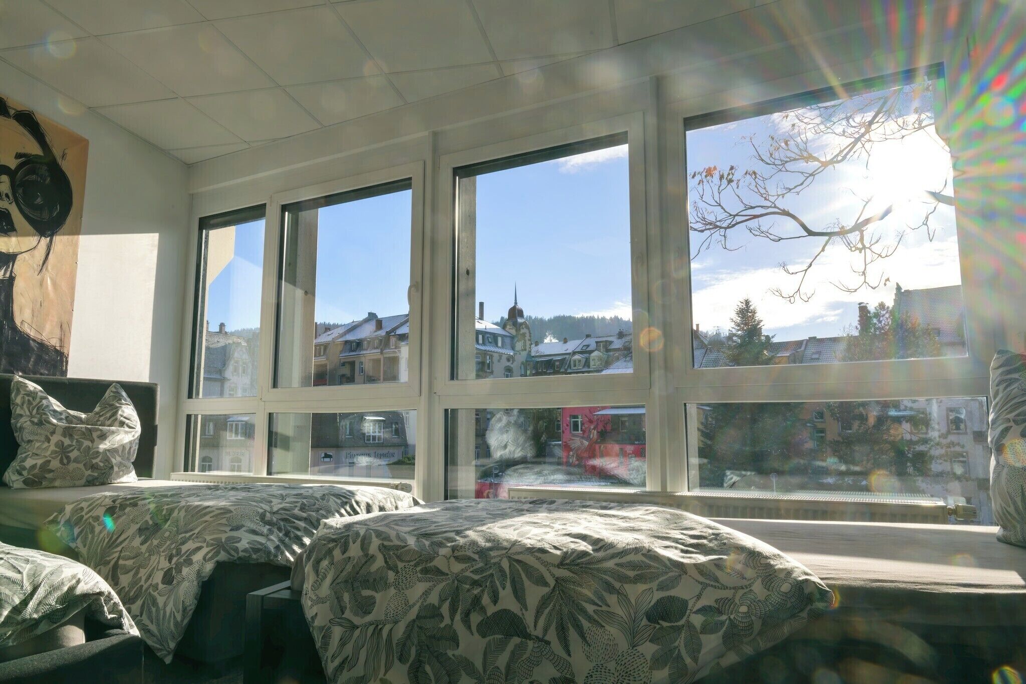 A room with two beds, large windows, and a view of a town.