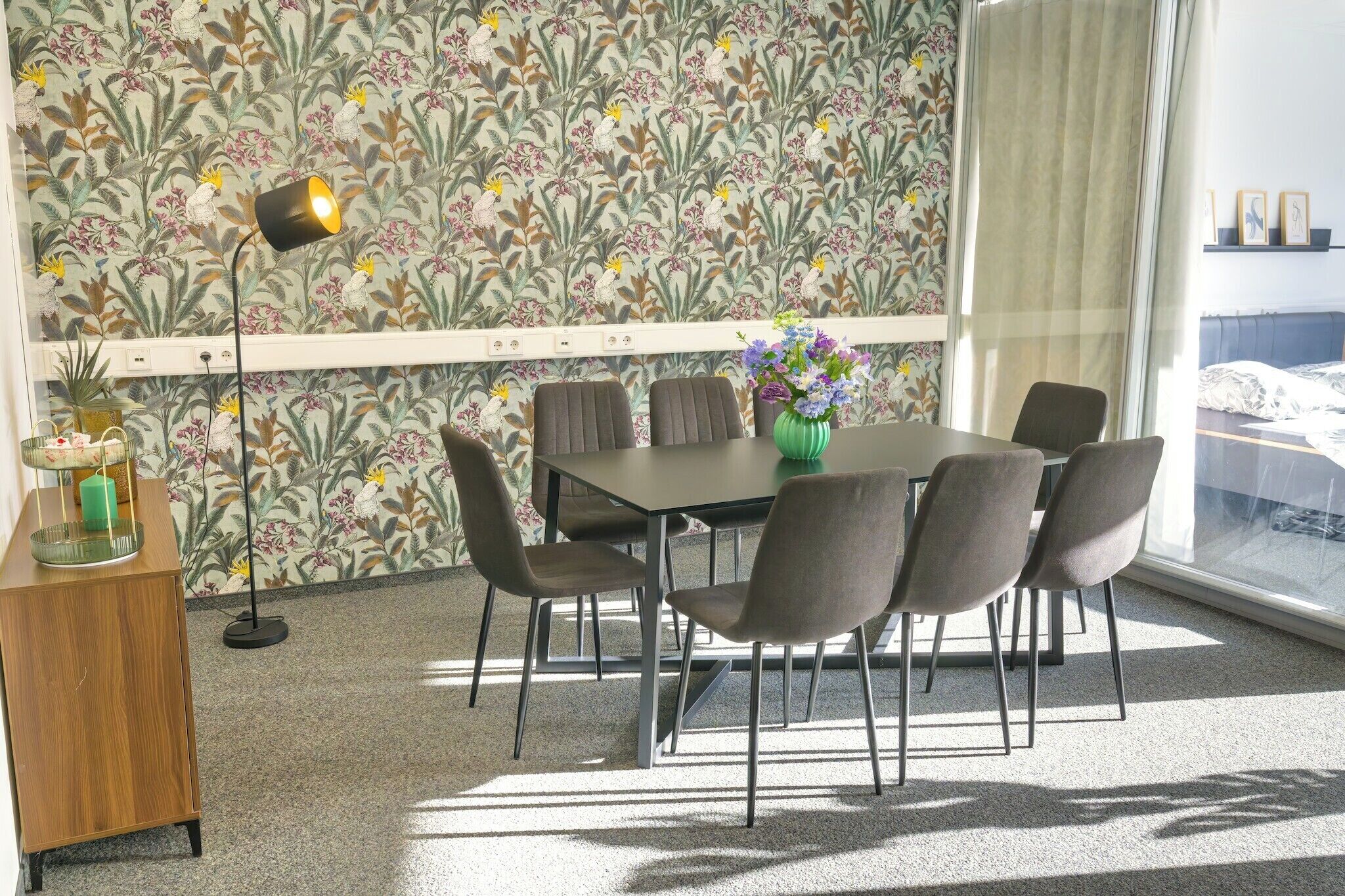 A dining area with a table and chairs, a vase with flowers, and a wall with a floral pattern.