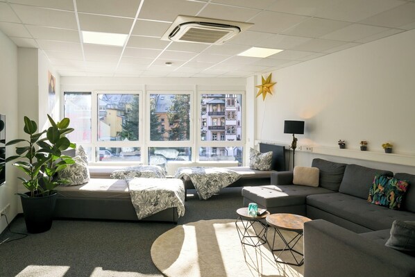 Ein modernes Wohnzimmer mit einer Couch, einem Couchtisch und einem Fernseher. Es gibt Fenster mit Blick auf Gebäude.