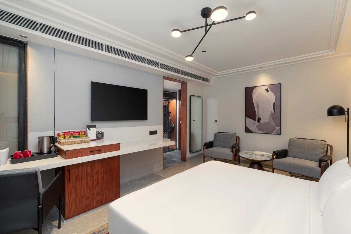 A hotel room with a bed, a desk with a coffee maker, a flat-screen TV, a chair, and a painting on the wall.