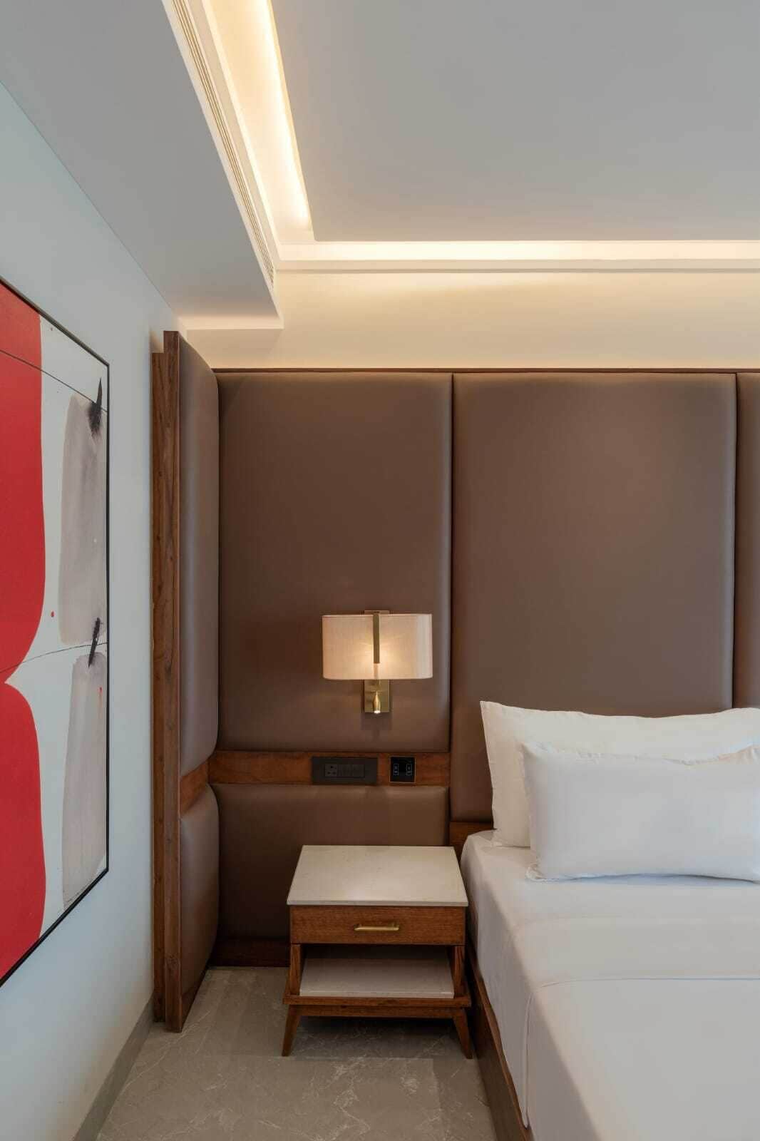 A hotel room with a bed, bedside table, lamp, and artwork on the wall.