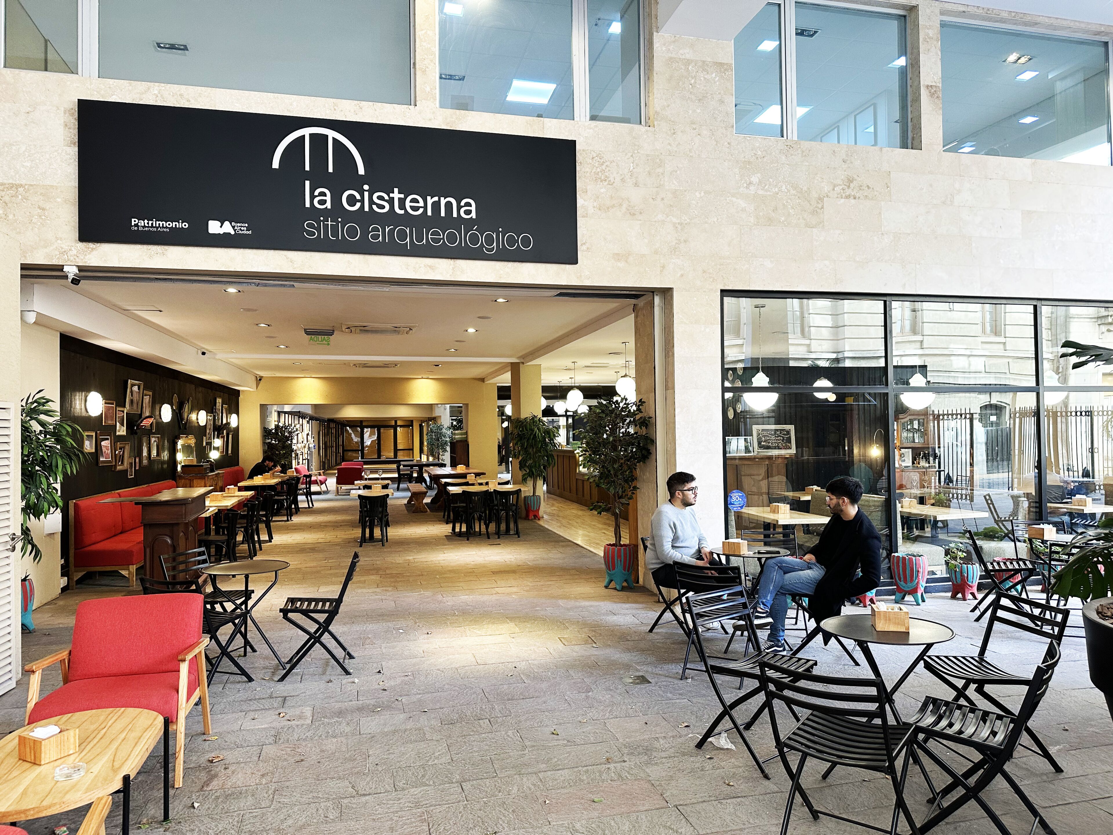 An indoor café with tables and chairs, a red bench, and a sign reading 'la cisterna sitio arqueológico'.