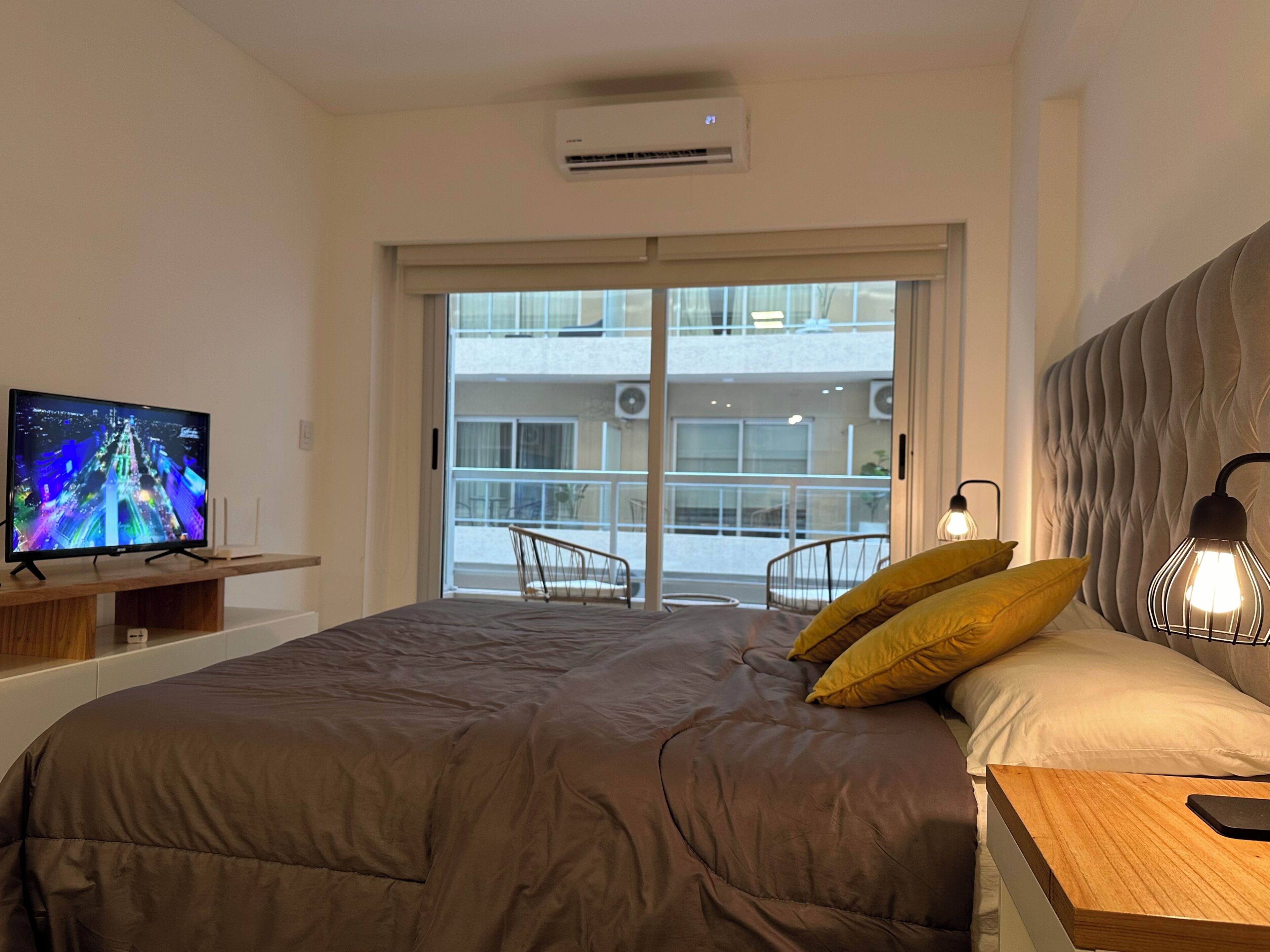 A bedroom with a bed, a TV on a stand, a bedside table, and a view of a balcony with chairs.