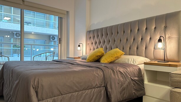A modern bedroom with a large bed, two bedside lamps, and a view of the outside balcony. - Vrbo Property (Buenos Aires)
