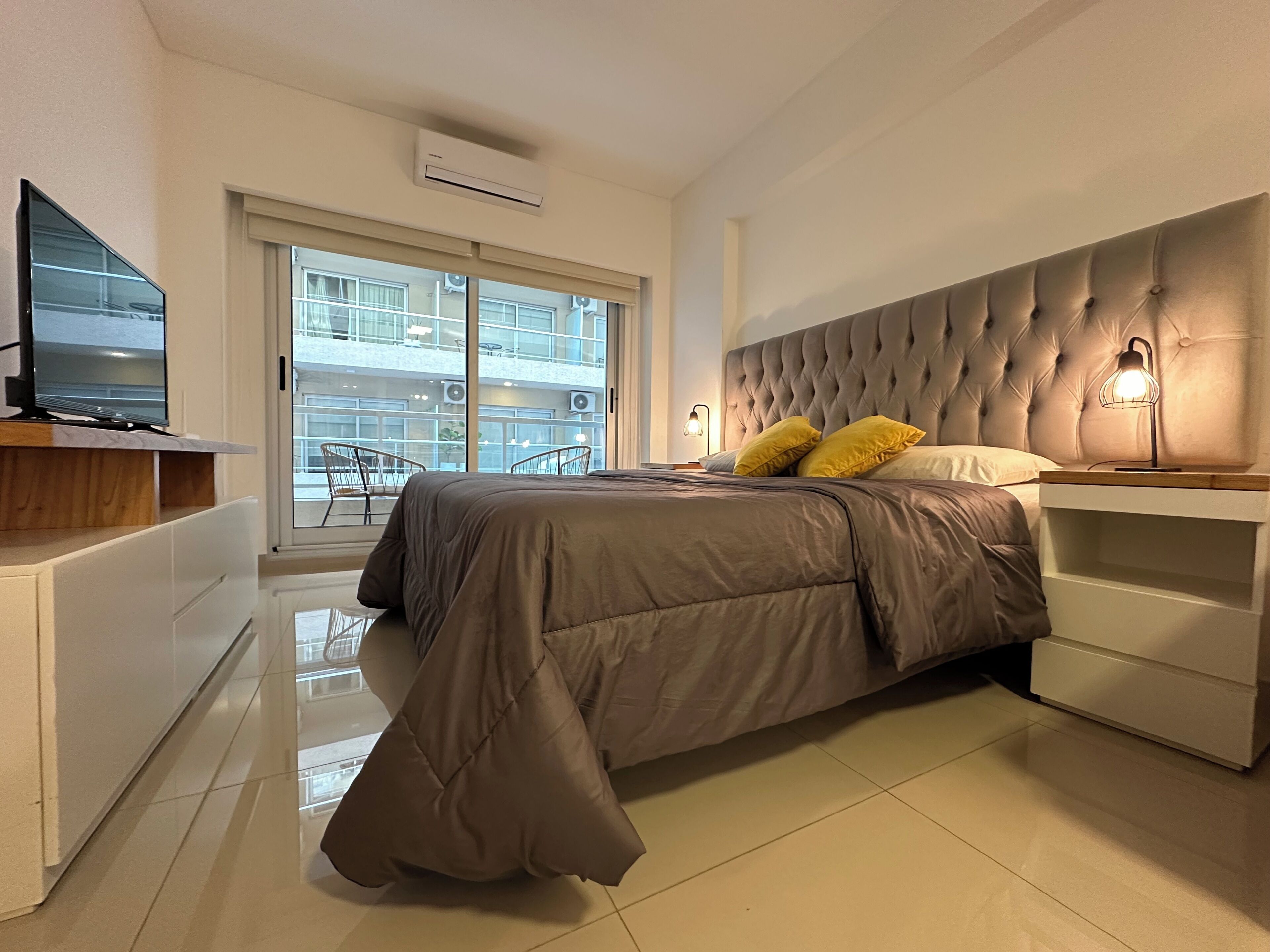 Un dormitorio moderno con una cama grande, mesitas de noche, un televisor y vistas al balcón exterior.