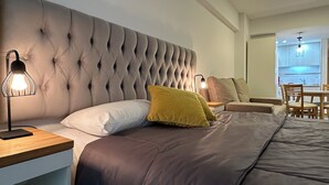 A bedroom with a tufted headboard, a bedside table with a lamp, a bed with a grey and yellow pillow, and a view of a kitchen area. - Vrbo Property (Buenos Aires)