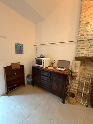 A room with a tiled floor, a wooden cabinet with drawers, a small trash bin, a wall-mounted shelf, and a basket rack. - Studio Zen- Éco Hameau au porte du marais poitevin (Liez)