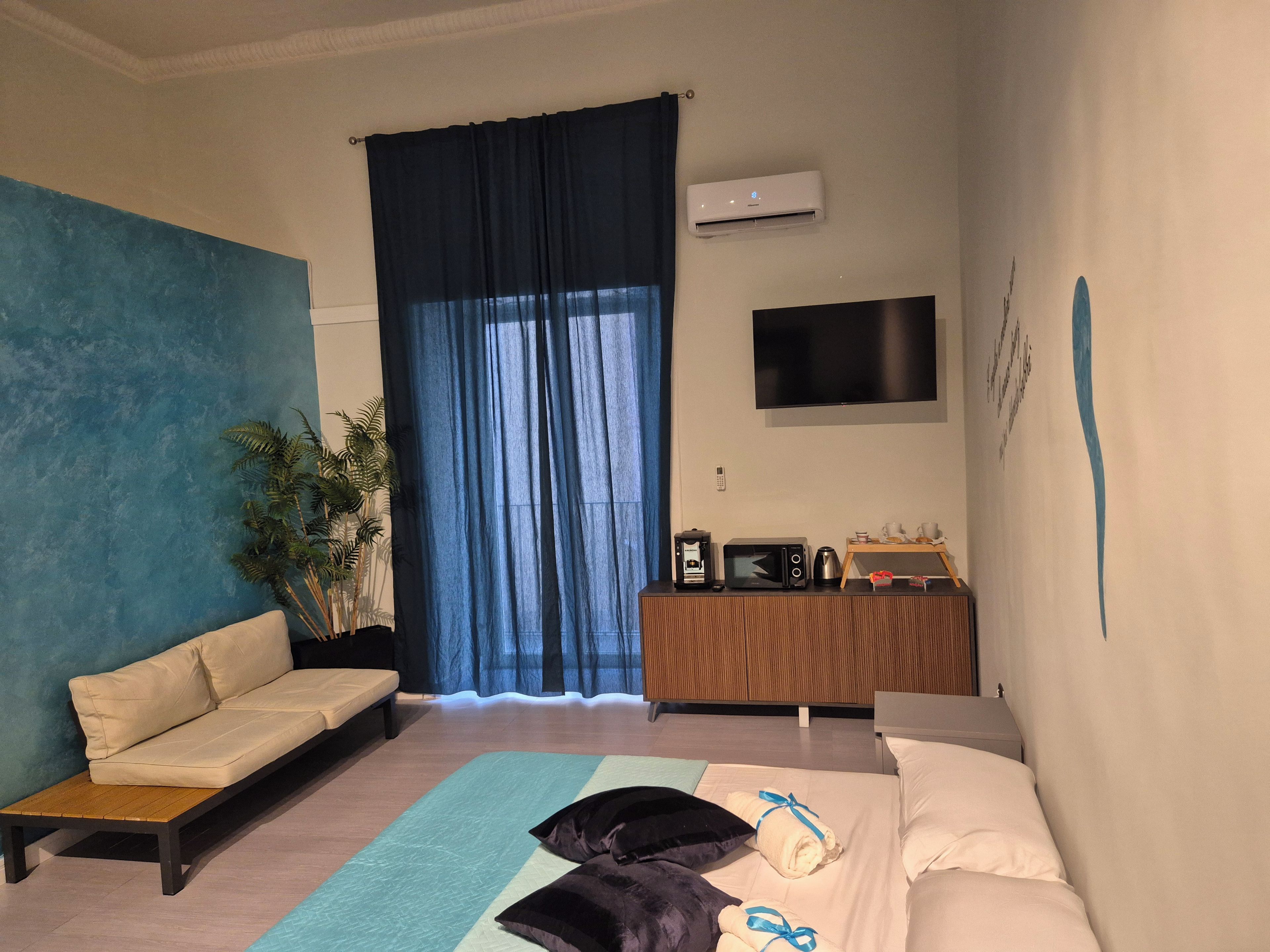 Una habitación de hotel moderna con sofá, cama, escritorio con computadora, televisor y un ventanal con cortinas.