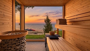 A wooden sauna room with a view of the sea at sunset, a bench, a wooden bucket, and a stack of towels. - Lakehouse 99 - Tranquil Waterfront Getaway in the heart of Prince Edward County (Prince Edward)