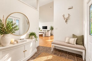 Una sala moderna con una consola blanca, un espejo redondo, un banco con cojines y una cabeza de venado decorativa.