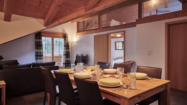 Zona pranzo con un tavolo di legno apparecchiato per quattro persone, circondato da sedie. Sullo sfondo, si vedono un divano e una televisione.