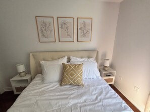 A neatly made bed with white bedding and a single patterned pillow, flanked by two bedside tables with lamps, in a room with a plain wall and three framed botanical prints. - Condo in the National Capital (Gatineau)