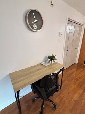 Quarto com escrivaninha de madeira, cadeira de escritório preta, uma pequena planta em vaso e um relógio de parede.