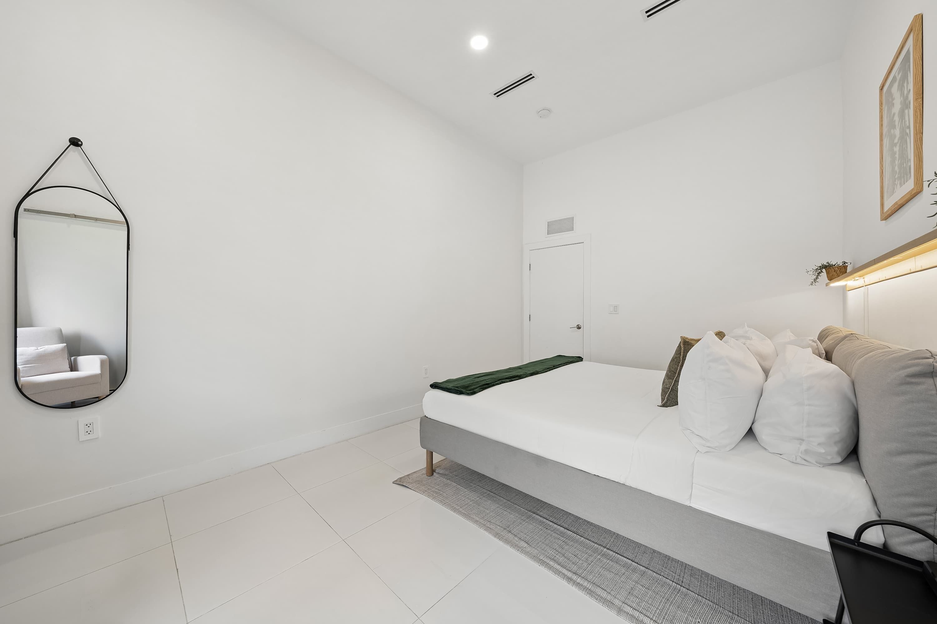 A minimalist bedroom with a white bed, a mirror, a door, and a small bedside table.