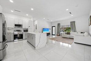 A modern kitchen with a central island, a dining area with a TV, and a living room with a sofa and a view of the outdoors. - Casa Azure Family Friendly Home with Heated Pool (Miami)