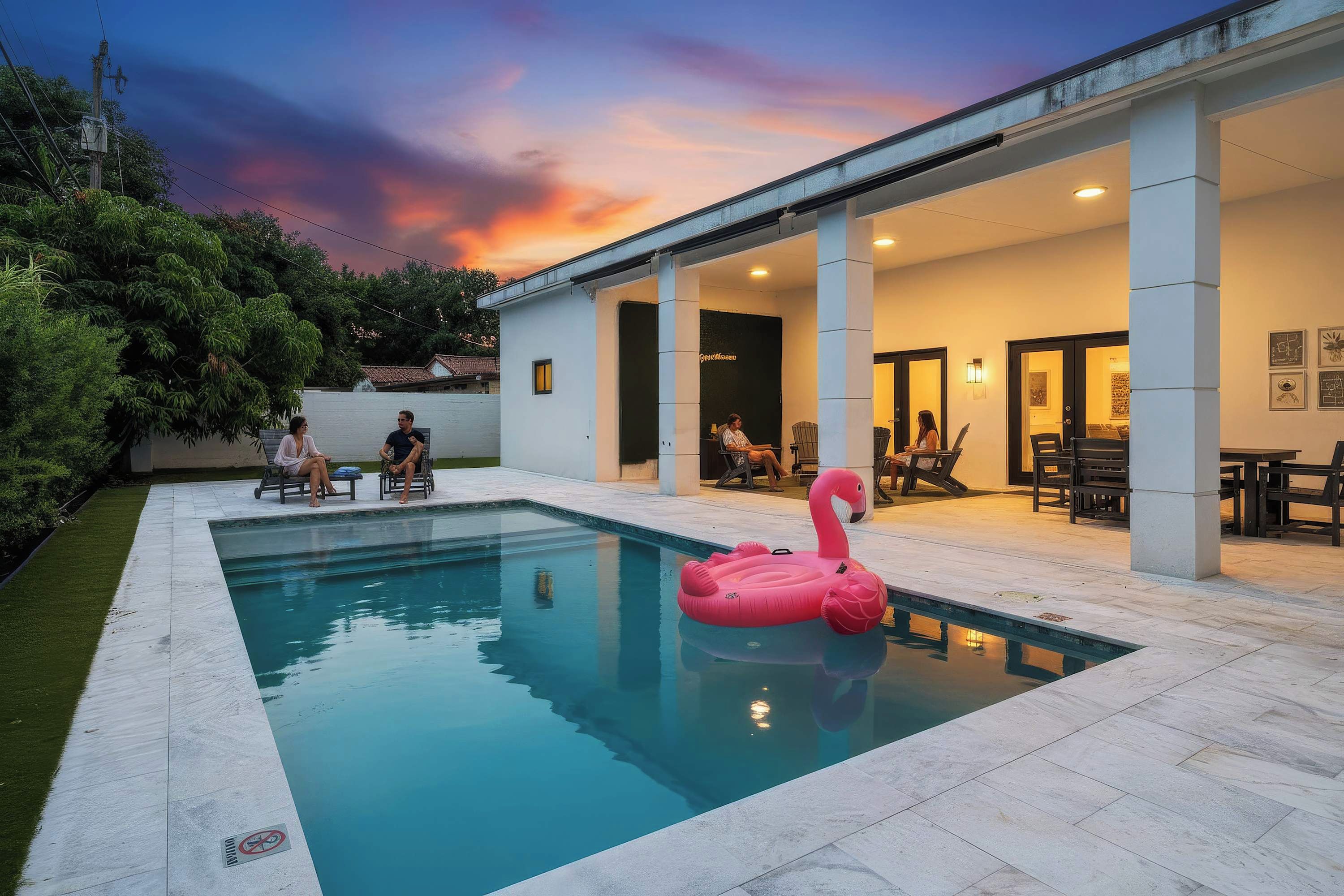 En swimmingpool med en lyserød flamingo-svømmebøje, omgivet af en overdækket terrasse med siddepladser.