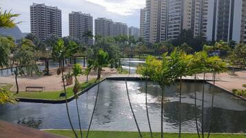 Área ajardinada com um lago ornamental, palmeiras e vista para edifícios altos ao fundo.