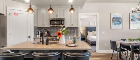 Una cocina moderna con una isla central, taburetes de bar, una zona de comedor con mesa y sillas, y un dormitorio con cama y mesita de noche.