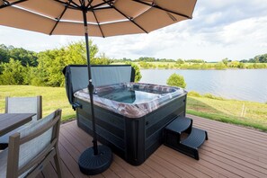 Uma banheira de hidromassagem em um deck de madeira, com guarda-sol, com vista para um lago.